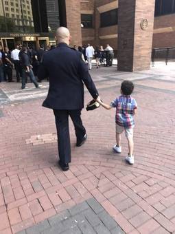 A uniformed officer and a small boy walking hand in hand, seen from behind