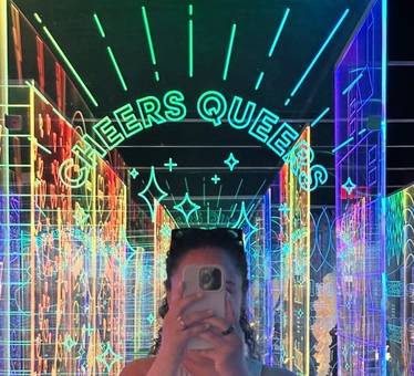 Jessenia taking a mirror selfie under a neon sign in a rainbow-lit mirrored room